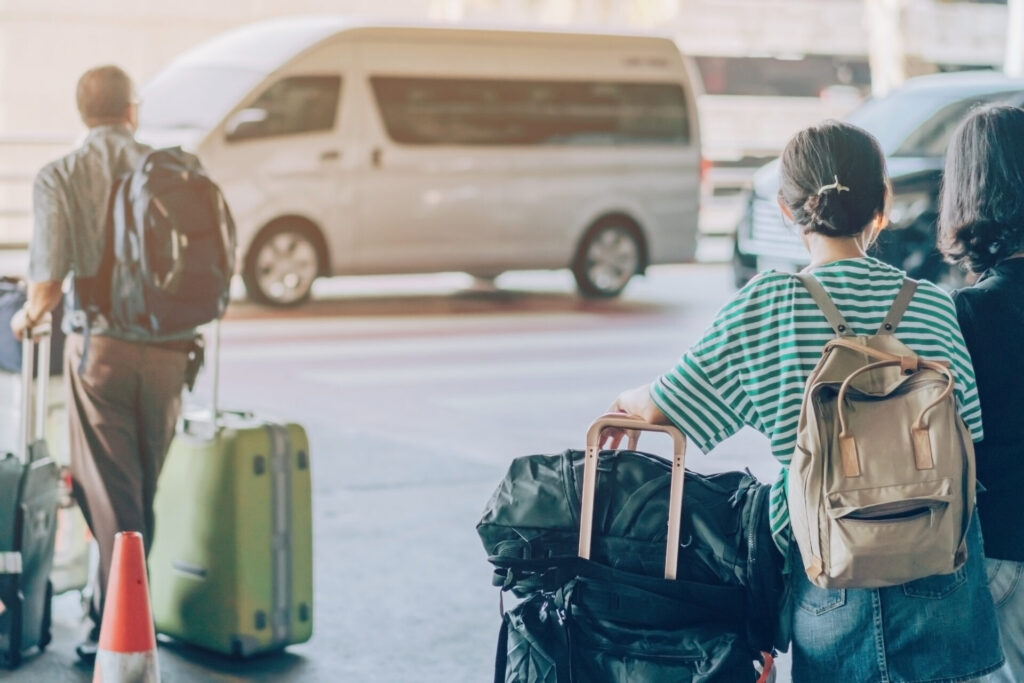 Passengers with big roller luggage stand to wait for the car to pick up at airport arrival terminal.
