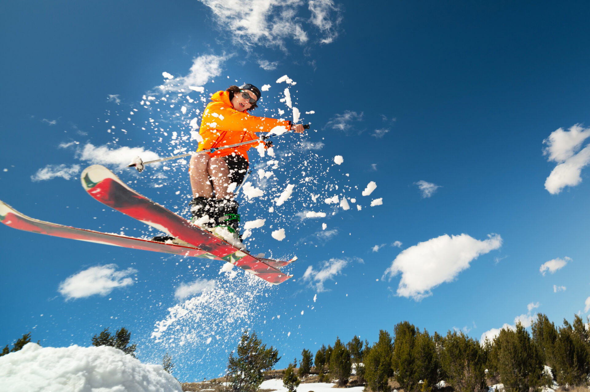 Man skier in flight after jumping from a kicker in the spring against the backdrop of mountains and blue sky.